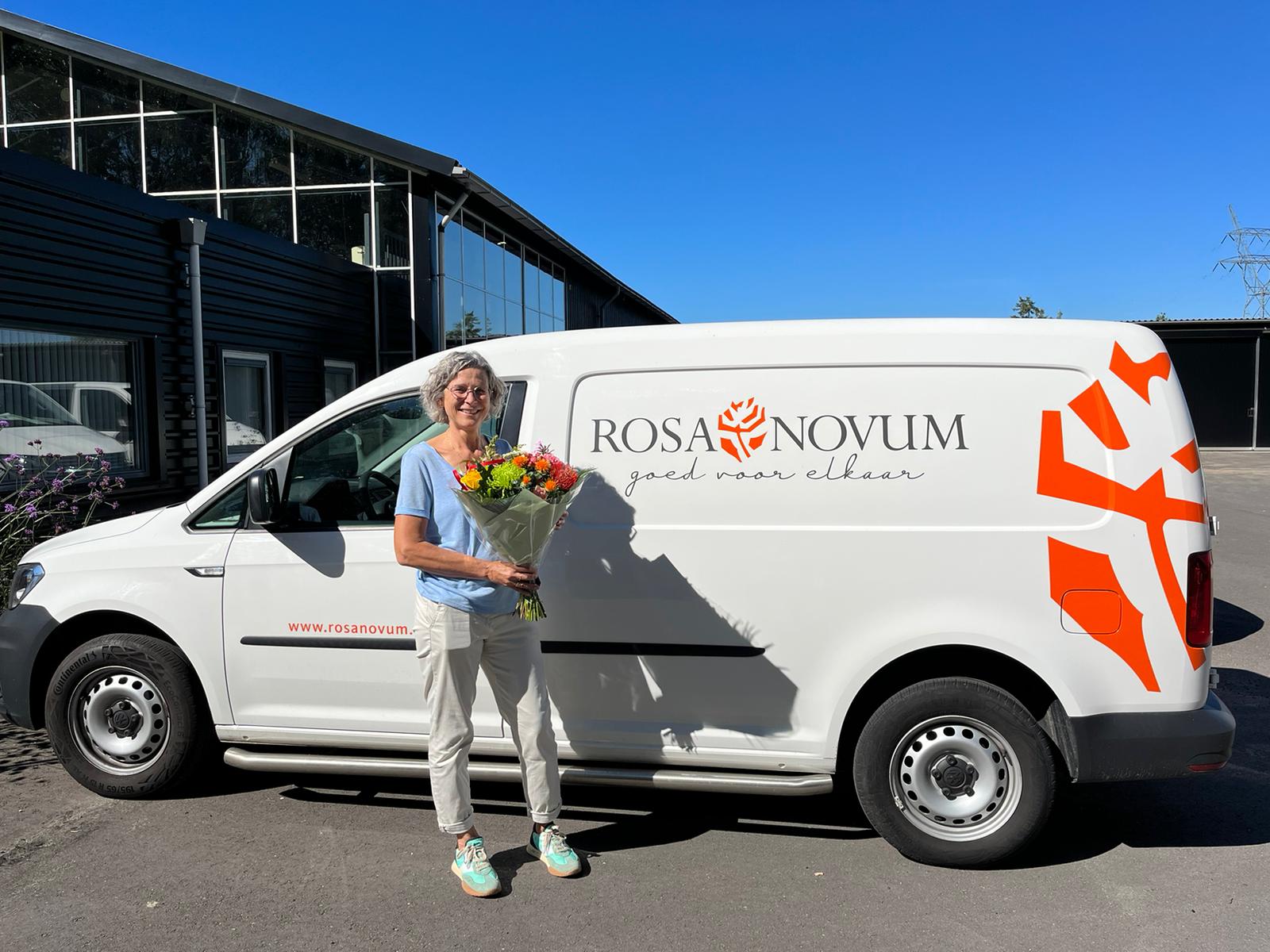 vrouw staat voor busje en ontvangt bloemen omdat zij genomineerd is voor de Hartstikke Sociaal Bokaal